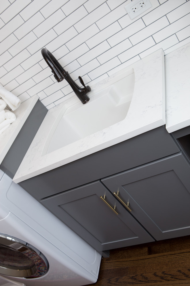 This is an example of a mid-sized transitional u-shaped utility room in Chicago with a farmhouse sink, shaker cabinets, quartz benchtops, white splashback, subway tile splashback, medium hardwood floors, brown floor, white benchtop, grey cabinets, beige walls and a side-by-side washer and dryer.