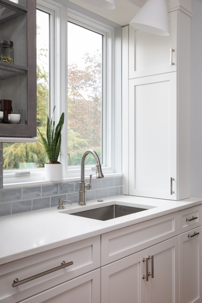 Example of a mid-sized transitional l-shaped medium tone wood floor and brown floor enclosed kitchen design with an undermount sink, shaker cabinets, white cabinets, quartz countertops, blue backsplash, subway tile backsplash, stainless steel appliances, an island and white countertops