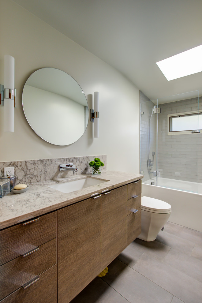 This is an example of a mid-sized contemporary master bathroom in San Francisco with flat-panel cabinets, light wood cabinets, a corner tub, a shower/bathtub combo, a one-piece toilet, gray tile, ceramic tile, white walls, ceramic floors, an undermount sink, engineered quartz benchtops, grey floor, a hinged shower door and beige benchtops.