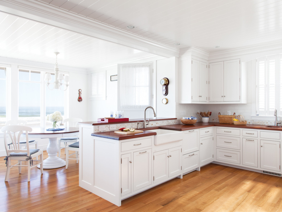 This is an example of a beach style u-shaped open plan kitchen in Portland Maine with a farmhouse sink, beaded inset cabinets, white cabinets, wood benchtops, white splashback, medium hardwood floors, a peninsula, brown floor, brown benchtop and timber.