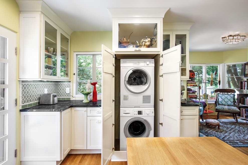 Mid-sized eclectic l-shaped laundry room in Orange County with a farmhouse sink, shaker cabinets, white cabinets, soapstone benchtops, medium hardwood floors and black benchtop.