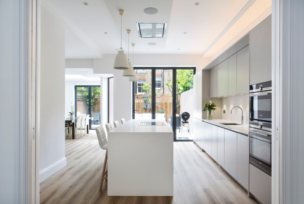 This is an example of a mid-sized transitional eat-in kitchen in London with an undermount sink, flat-panel cabinets, white cabinets, quartzite benchtops, stainless steel appliances, light hardwood floors, with island, beige floor and white benchtop.