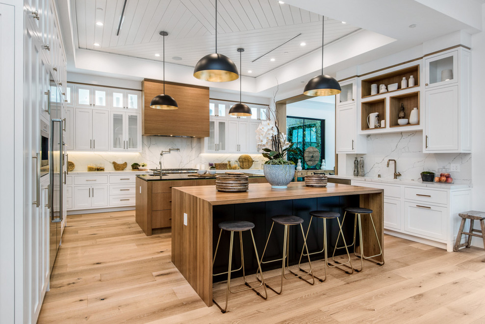 This is an example of a transitional u-shaped kitchen in Los Angeles with shaker cabinets, white cabinets, white splashback, stone slab splashback, stainless steel appliances, light hardwood floors, multiple islands, beige floor and white benchtop.