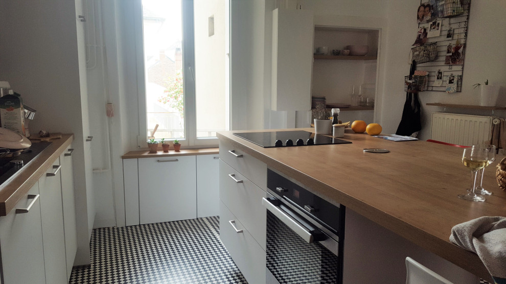 This is an example of a mid-sized transitional galley open plan kitchen in Paris with an undermount sink, flat-panel cabinets, white cabinets, wood benchtops, white splashback, subway tile splashback, panelled appliances, linoleum floors, with island, multi-coloured floor and brown benchtop.