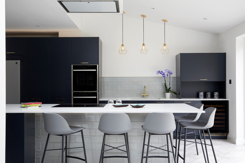 Photo of a contemporary open plan kitchen in Kent with a drop-in sink, flat-panel cabinets, blue cabinets, solid surface benchtops, metallic splashback, glass tile splashback, black appliances, porcelain floors, with island, grey floor and white benchtop.