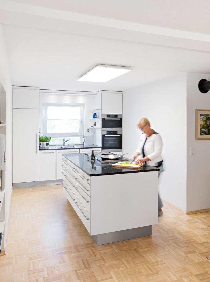 Mid-sized modern single-wall open plan kitchen in Stuttgart with flat-panel cabinets, white cabinets, granite benchtops, with island and black benchtop.