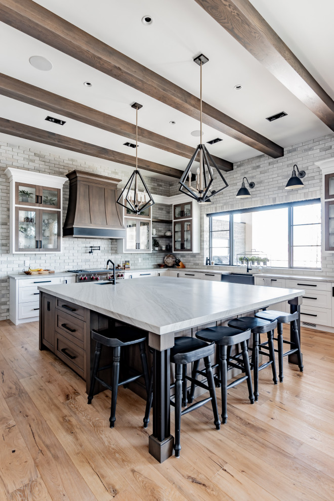 Large transitional l-shaped medium tone wood floor, brown floor and exposed beam eat-in kitchen photo in Calgary with shaker cabinets, white cabinets, granite countertops, gray backsplash, brick backsplash, an island, gray countertops and stainless steel appliances