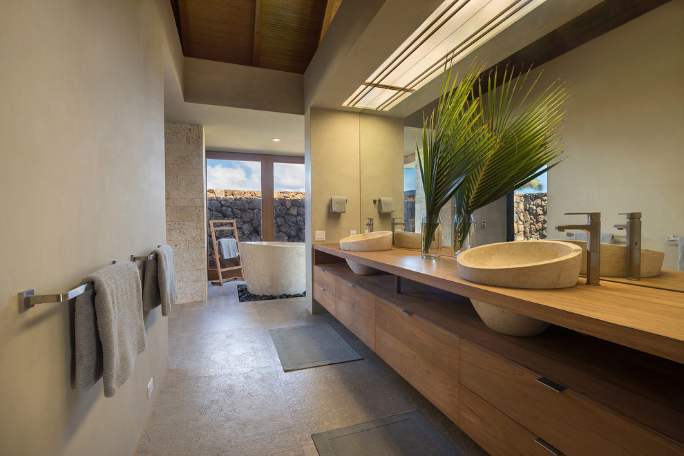 Design ideas for a tropical master bathroom in Hawaii with medium wood cabinets, a freestanding tub, limestone floors, a vessel sink, wood benchtops, grey floor, flat-panel cabinets and beige walls.