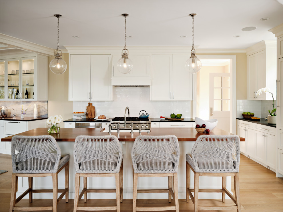 Photo of a beach style l-shaped kitchen in Boston with light hardwood floors, with island, shaker cabinets, white cabinets, white splashback, subway tile splashback, stainless steel appliances and black benchtop.