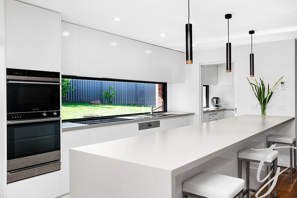 Photo of a large contemporary galley kitchen pantry in Melbourne with a double-bowl sink, white cabinets, solid surface benchtops, timber splashback, stainless steel appliances, light hardwood floors, with island and white benchtop.