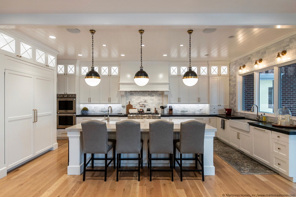 Large traditional u-shaped open plan kitchen in Salt Lake City with a farmhouse sink, recessed-panel cabinets, white cabinets, granite benchtops, grey splashback, porcelain splashback, stainless steel appliances, medium hardwood floors, with island, brown floor, black benchtop and timber.
