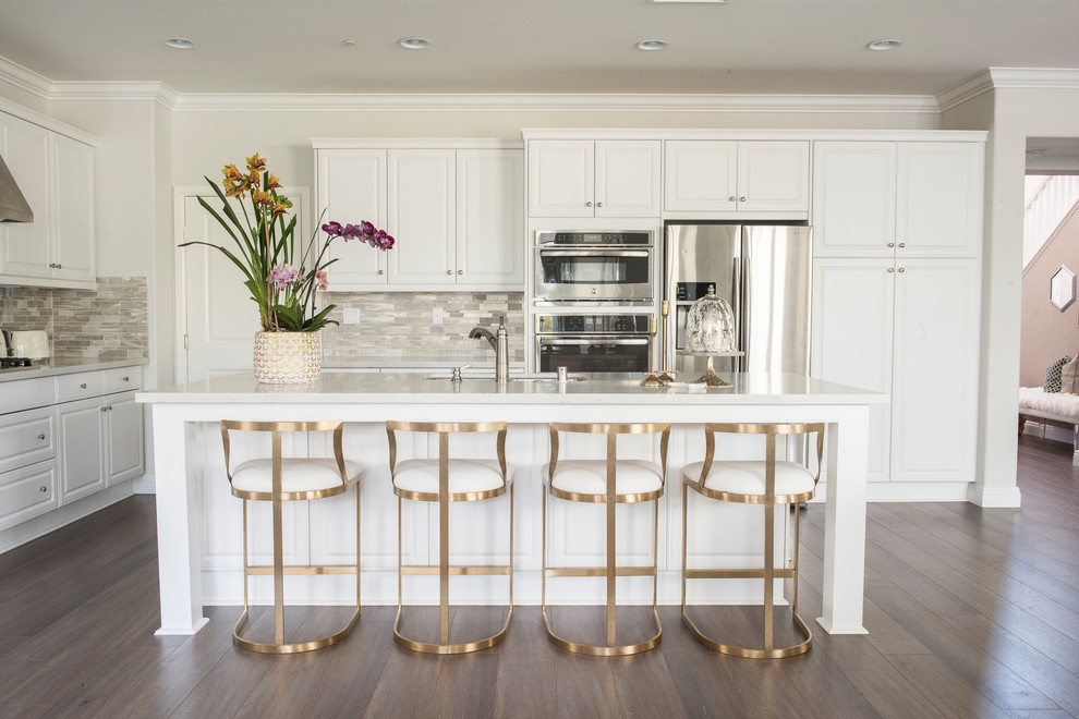 Example of a mid-sized transitional l-shaped brown floor and dark wood floor open concept kitchen design with an undermount sink, raised-panel cabinets, white cabinets, quartz countertops, metallic backsplash, stainless steel appliances, an island and white countertops