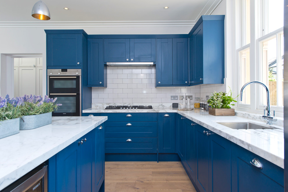 Inspiration for a large transitional l-shaped open plan kitchen in London with shaker cabinets, blue cabinets, marble benchtops, white splashback, ceramic splashback, stainless steel appliances, light hardwood floors, with island, brown floor and an undermount sink.