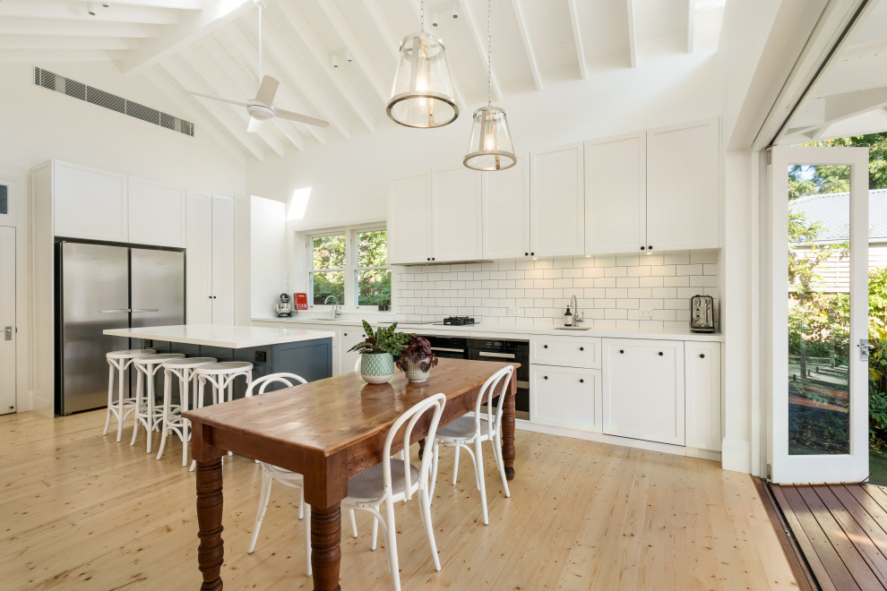 Transitional l-shaped eat-in kitchen in Sydney with an undermount sink, shaker cabinets, white cabinets, white splashback, subway tile splashback, black appliances, light hardwood floors, with island, beige floor, white benchtop, exposed beam and vaulted.