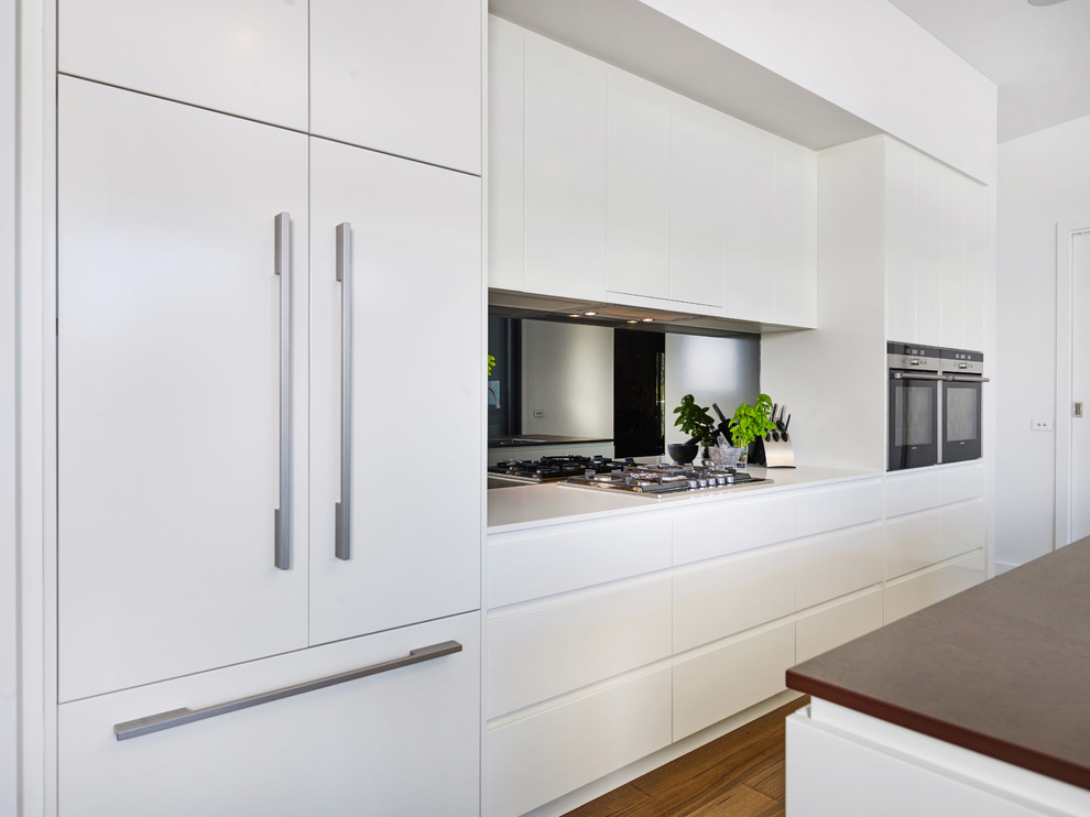 Inspiration for a large contemporary galley kitchen pantry in Other with a drop-in sink, flat-panel cabinets, white cabinets, solid surface benchtops, metallic splashback, mirror splashback, stainless steel appliances, light hardwood floors, with island, brown floor and brown benchtop.