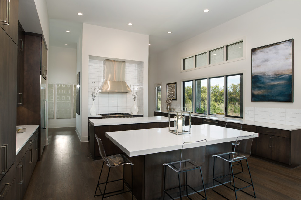 This is an example of a large contemporary single-wall open plan kitchen in Wichita with flat-panel cabinets, dark wood cabinets, multiple islands, solid surface benchtops, white splashback, porcelain splashback, stainless steel appliances, an undermount sink and dark hardwood floors.