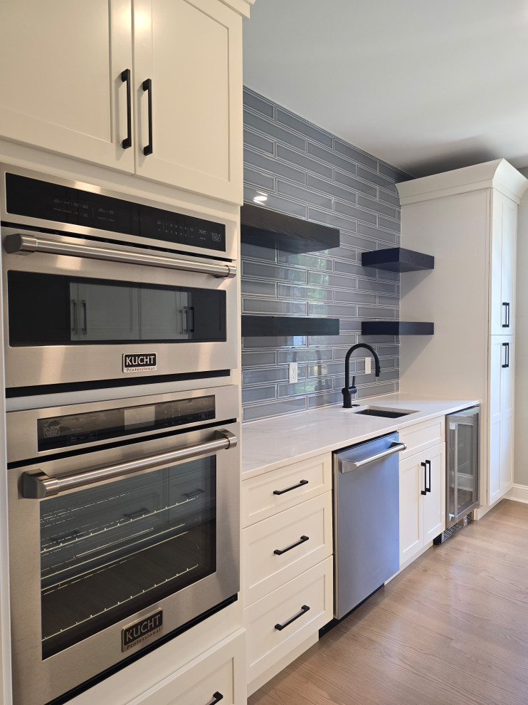 Example of a large minimalist l-shaped medium tone wood floor and brown floor kitchen design with shaker cabinets, white cabinets, quartz countertops, gray backsplash, subway tile backsplash, stainless steel appliances and white countertops