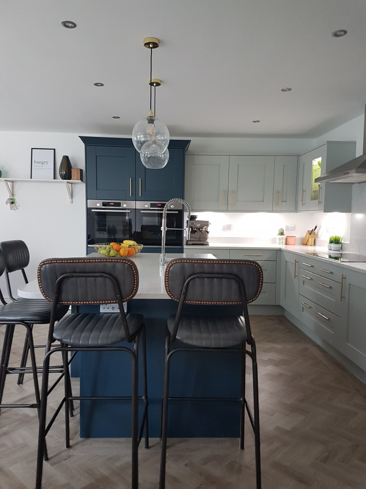 Mid-sized contemporary l-shaped open plan kitchen in West Midlands with an integrated sink, shaker cabinets, grey cabinets, quartzite benchtops, grey splashback, ceramic splashback, black appliances, medium hardwood floors, with island, brown floor and grey benchtop.