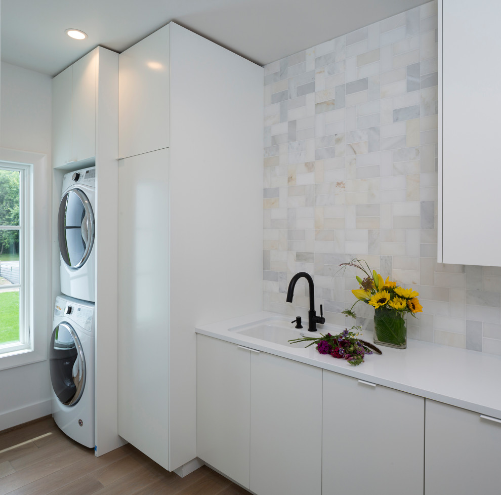 Photo of a contemporary separated utility room in Minneapolis with a single-bowl sink, flat-panel cabinets, white cabinets, multi-coloured splashback, white walls, dark hardwood flooring, a stacked washer and dryer and white worktops.