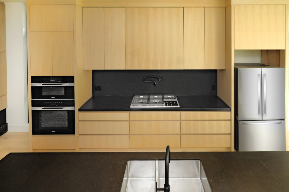 バンクーバーにあるラグジュアリーな広いコンテンポラリースタイルのおしゃれなキッチン (ドロップインシンク、フラットパネル扉のキャビネット、ベージュのキャビネット、クオーツストーンカウンター、黒いキッチンパネル、セメントタイルのキッチンパネル、黒い調理設備、淡色無垢フローリング、ベージュの床、黒いキッチンカウンター、三角天井) の写真