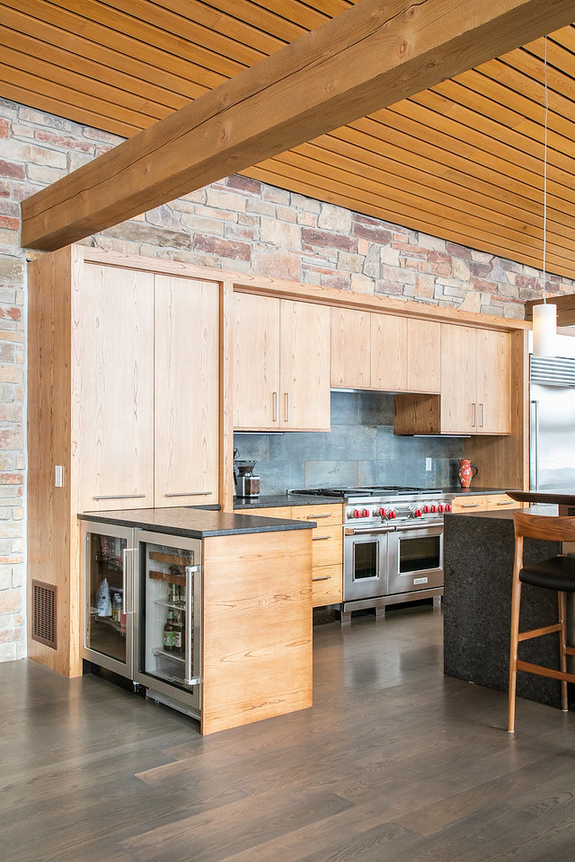 Large mountain style l-shaped medium tone wood floor and brown floor open concept kitchen photo with flat-panel cabinets, light wood cabinets, granite countertops, black backsplash, stainless steel appliances, an island, black countertops, a double-bowl sink and porcelain backsplash