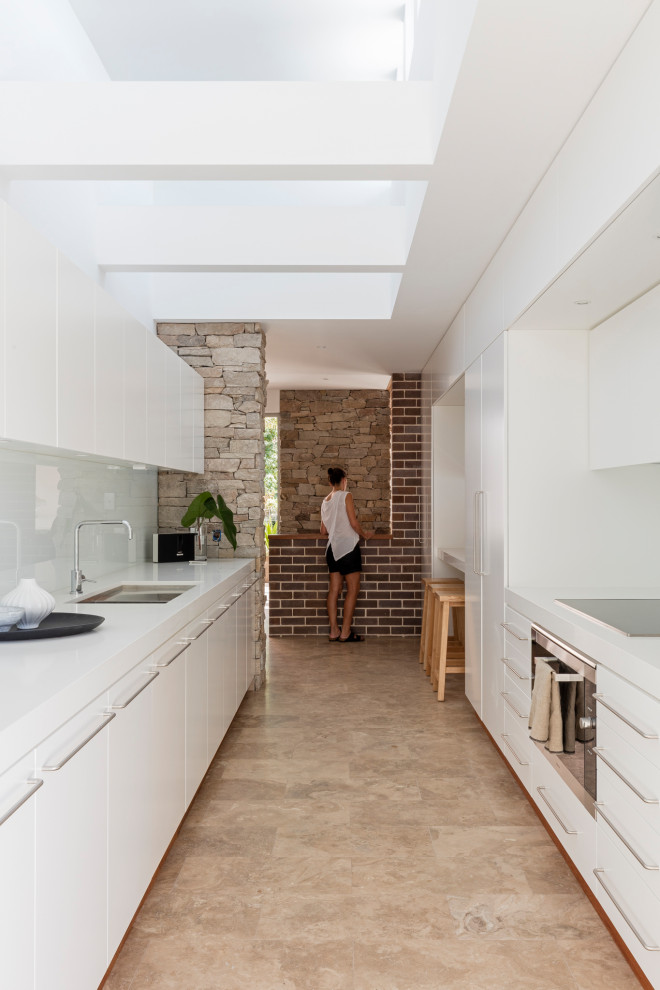Inspiration for a large midcentury galley separate kitchen in Sydney with an undermount sink, quartz benchtops, white splashback, glass sheet splashback, black appliances, travertine floors, brown floor and white benchtop.