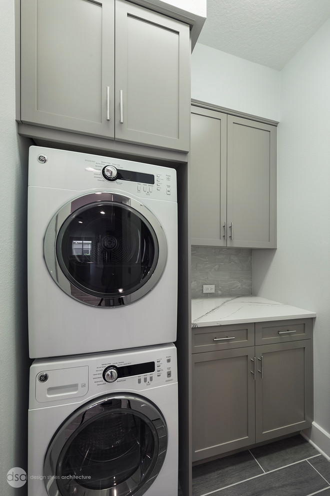 Design ideas for a large transitional single-wall dedicated laundry room in Tampa with shaker cabinets, grey cabinets, quartz benchtops, white walls, porcelain floors, a stacked washer and dryer and grey floor.