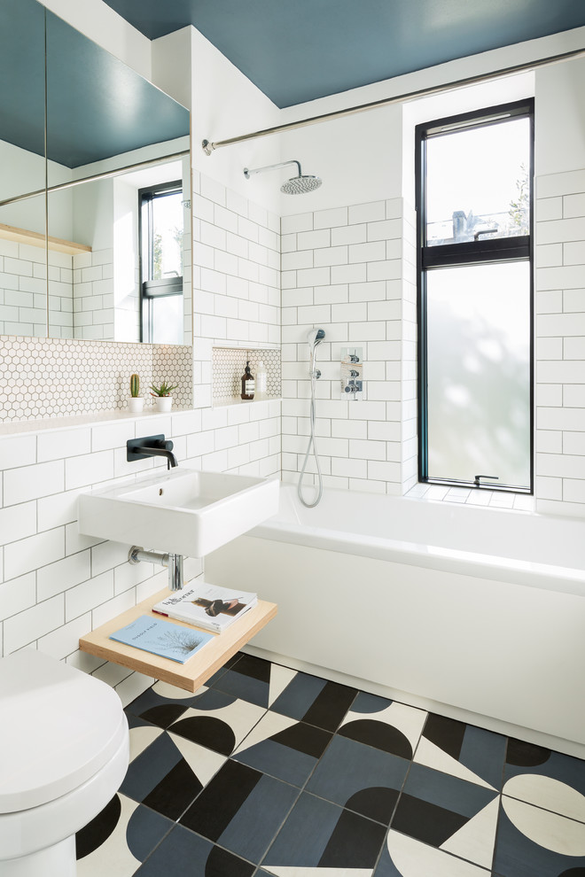This is an example of a mid-sized scandinavian bathroom in London with a drop-in tub, white tile, subway tile, white walls, ceramic floors, a wall-mount sink, multi-coloured floor, a shower/bathtub combo and a wall-mount toilet.