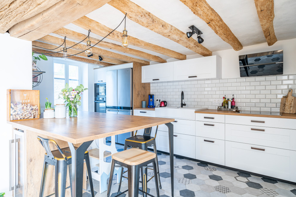 Photo of a mid-sized country single-wall open plan kitchen in Angers with a farmhouse sink, beaded inset cabinets, white cabinets, wood benchtops, white splashback, subway tile splashback, panelled appliances, ceramic floors, a peninsula, multi-coloured floor, brown benchtop and exposed beam.