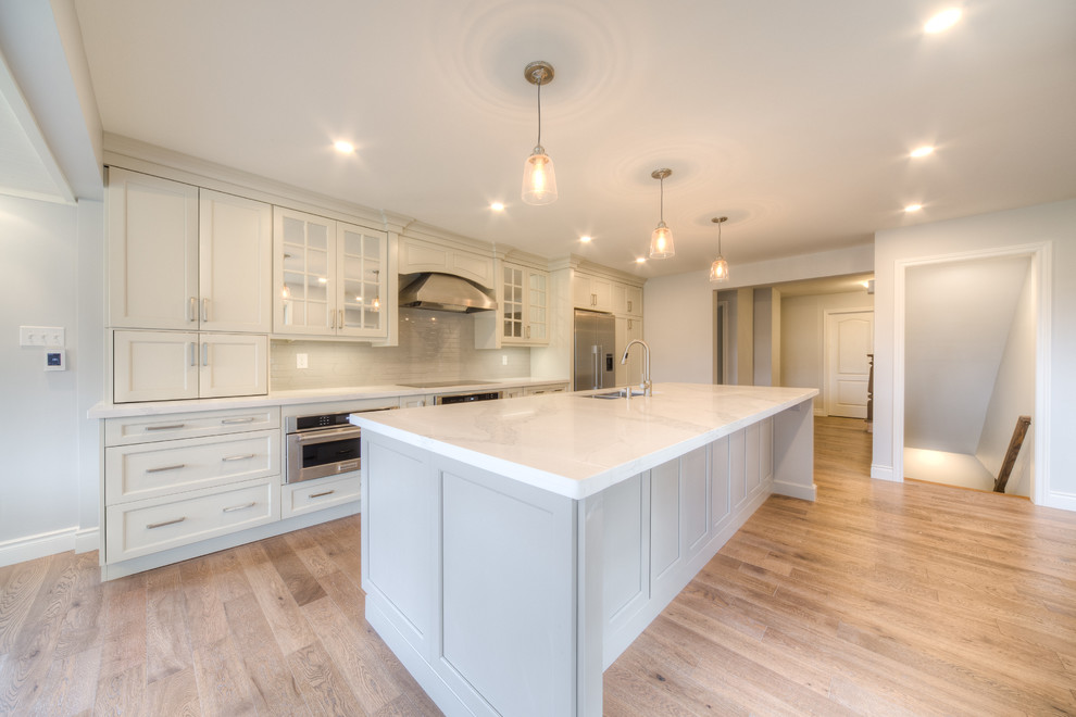 Mid-sized transitional galley light wood floor and beige floor open concept kitchen photo in Toronto with a double-bowl sink, recessed-panel cabinets, white cabinets, solid surface countertops, white backsplash, porcelain backsplash, stainless steel appliances, an island and white countertops