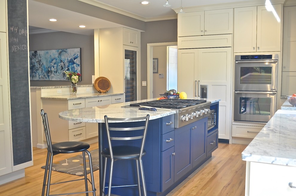 Example of a mid-sized transitional l-shaped light wood floor and brown floor enclosed kitchen design in Denver with a farmhouse sink, shaker cabinets, white cabinets, marble countertops, stainless steel appliances, an island, white backsplash and ceramic backsplash