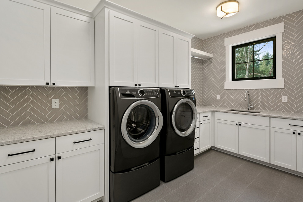 This is an example of a large country galley dedicated laundry room in Seattle with a drop-in sink, recessed-panel cabinets, white cabinets, quartz benchtops, grey splashback, ceramic splashback, white walls, ceramic floors, a stacked washer and dryer, grey floor and white benchtop.