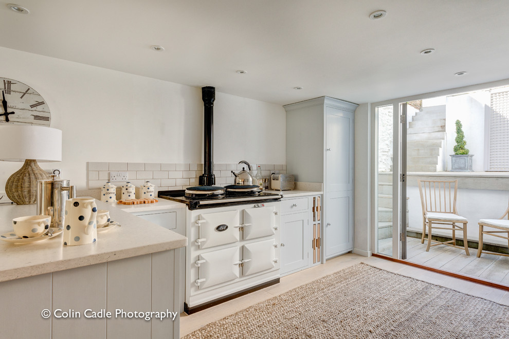 Small beach style u-shaped eat-in kitchen in Devon with a farmhouse sink, recessed-panel cabinets, distressed cabinets, marble benchtops, white splashback, ceramic splashback, white appliances and light hardwood floors.