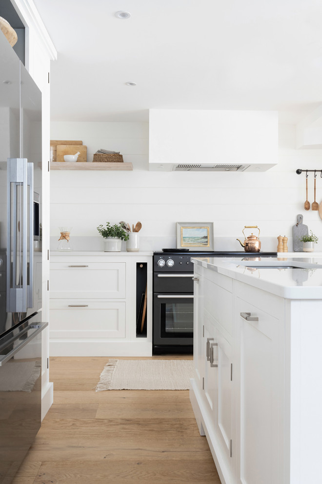 Photo of a coastal l-shaped kitchen in Devon with a submerged sink, shaker cabinets, white cabinets, white splashback, tonge and groove splashback, black appliances, light hardwood flooring, an island, beige floors and white worktops.