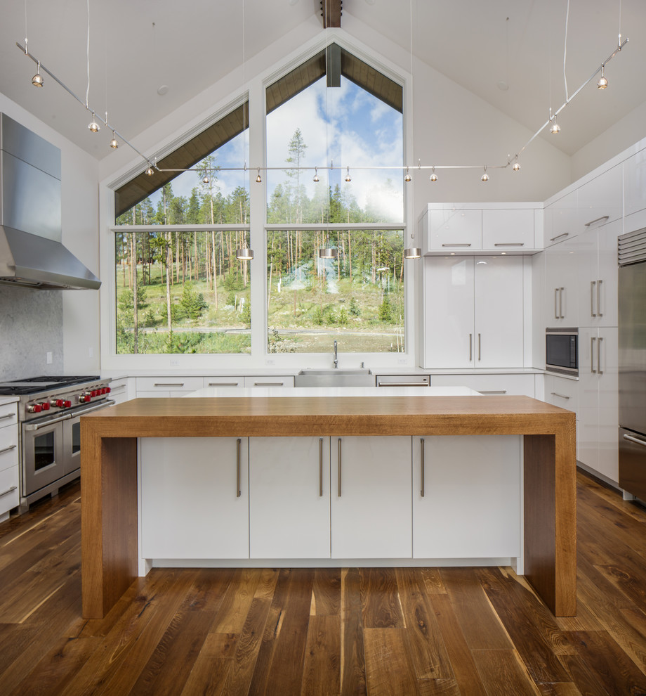 Design ideas for a contemporary kitchen in Denver with a farmhouse sink, white cabinets, stainless steel appliances, medium hardwood floors, flat-panel cabinets, wood benchtops, window splashback and multiple islands.