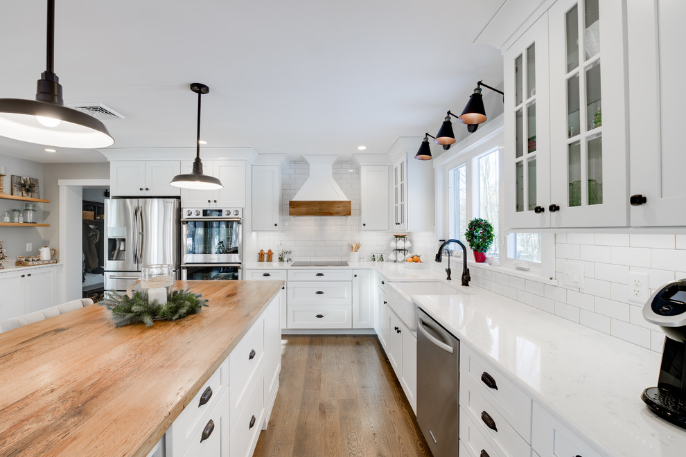 Country l-shaped kitchen in Philadelphia with a farmhouse sink, shaker cabinets, white cabinets, white splashback, subway tile splashback, stainless steel appliances, medium hardwood floors, with island, brown floor and white benchtop.
