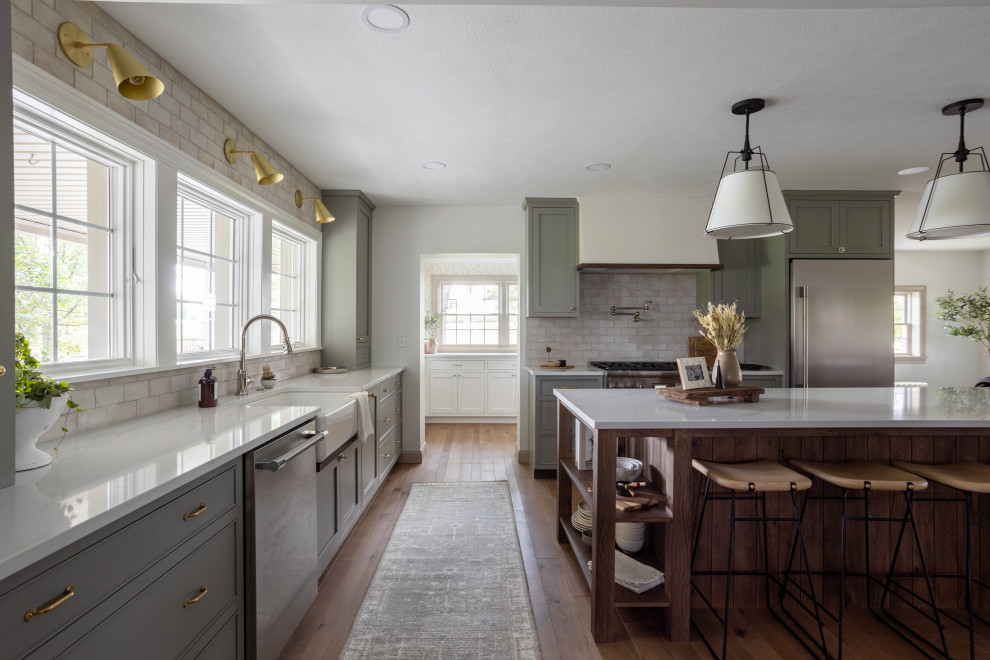 Photo of a galley eat-in kitchen in Cleveland with a farmhouse sink, shaker cabinets, green cabinets, white splashback, stone tile splashback, stainless steel appliances, medium hardwood floors, with island, brown floor and white benchtop.