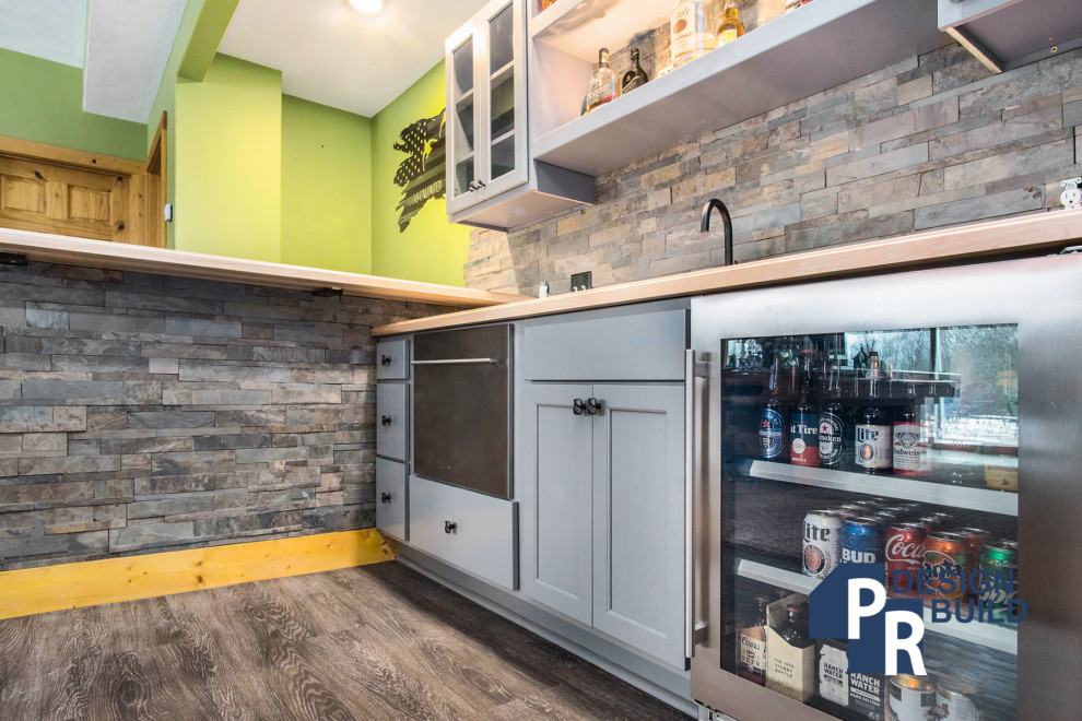 Medium sized rustic l-shaped wet bar in Grand Rapids with glass-front cabinets, grey cabinets, wood worktops, multi-coloured splashback, stone tiled splashback and brown worktops.