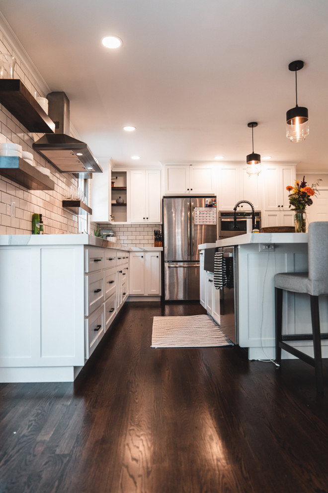 Design ideas for a large country l-shaped eat-in kitchen in Other with a farmhouse sink, shaker cabinets, white cabinets, quartz benchtops, white splashback, subway tile splashback, stainless steel appliances, dark hardwood floors, with island, brown floor and white benchtop.