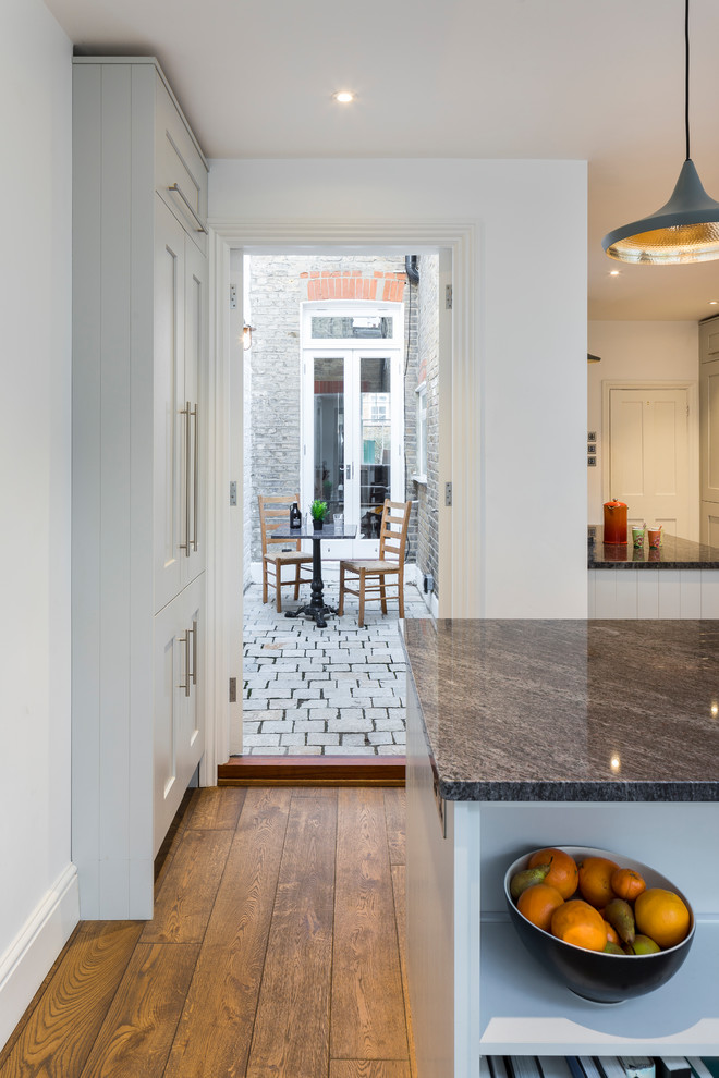 Large contemporary galley kitchen/diner in London with an integrated sink, shaker cabinets, grey cabinets, granite worktops, glass sheet splashback, black appliances, medium hardwood flooring, brown floors and an island.