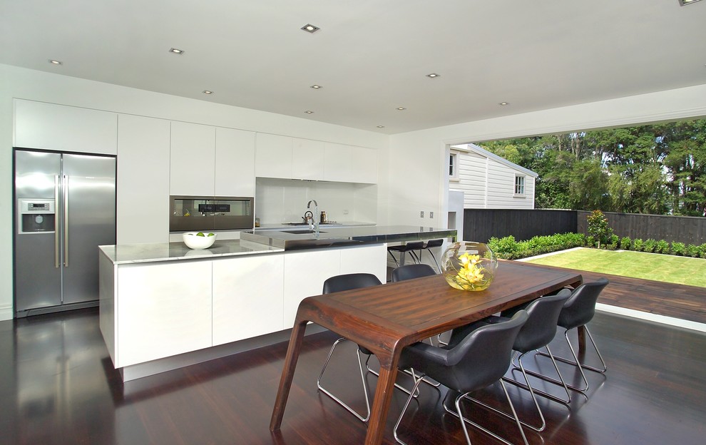 This is an example of an expansive contemporary l-shaped open plan kitchen in Auckland with stainless steel appliances, flat-panel cabinets, white cabinets, glass sheet splashback, with island, a single-bowl sink, marble benchtops, white splashback and dark hardwood floors.