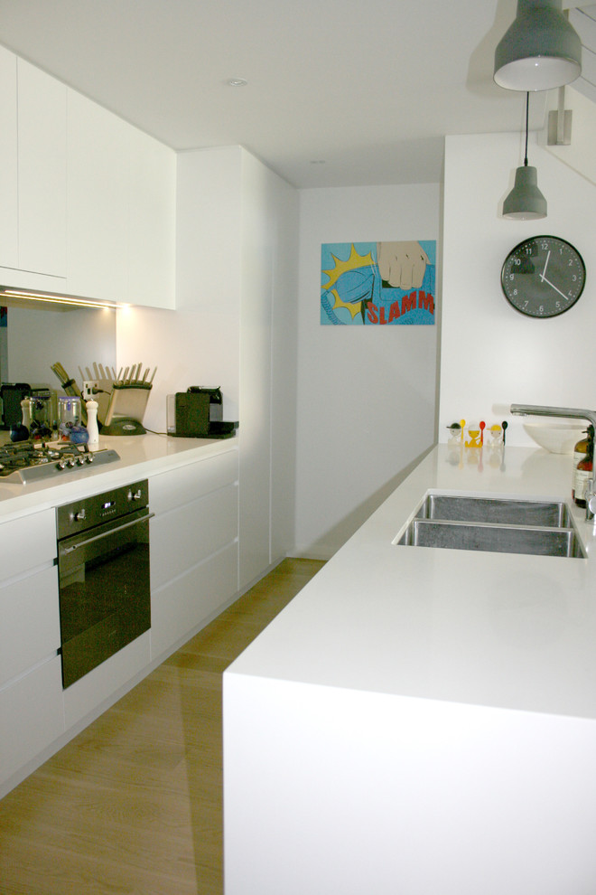 Mid-sized modern galley eat-in kitchen in Sydney with an undermount sink, white cabinets, solid surface benchtops, glass sheet splashback, stainless steel appliances, light hardwood floors and with island.
