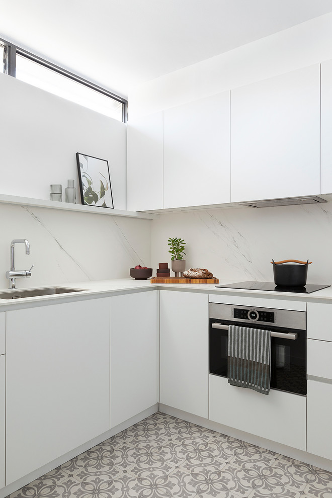 Contemporary l-shaped kitchen in Barcelona with an undermount sink, flat-panel cabinets, white cabinets, white splashback, stone slab splashback, stainless steel appliances, multi-coloured floor and white benchtop.