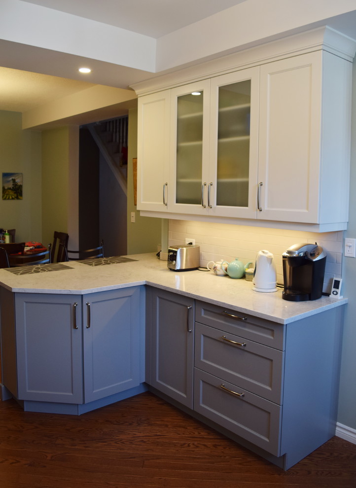 Example of a mid-sized transitional l-shaped medium tone wood floor eat-in kitchen design in Toronto with an undermount sink, shaker cabinets, white cabinets, quartz countertops, white backsplash, subway tile backsplash, stainless steel appliances and a peninsula