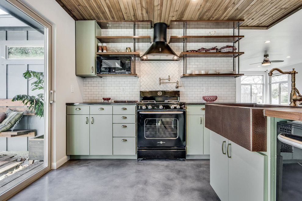 This is an example of an industrial l-shaped kitchen in Austin with a farmhouse sink, flat-panel cabinets, green cabinets, wood benchtops, white splashback, black appliances, concrete floors and grey floor.