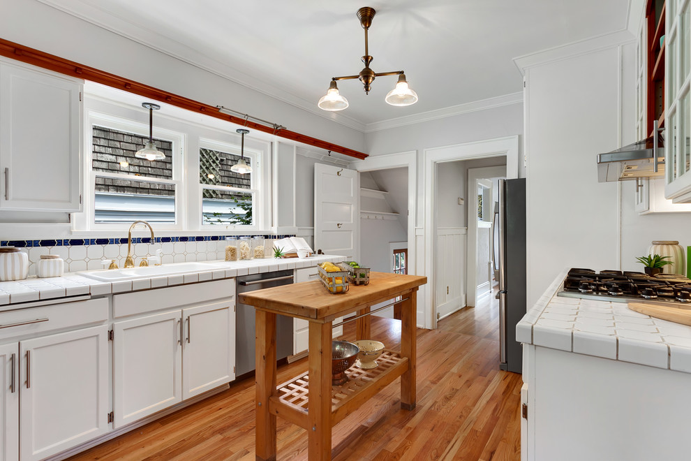 Photo of a mid-sized arts and crafts galley kitchen in Seattle with recessed-panel cabinets, white cabinets, white splashback, stainless steel appliances, dark hardwood floors, with island, brown floor and white benchtop.