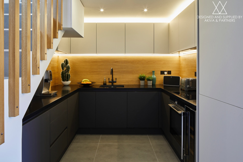 This is an example of a small industrial u-shaped open plan kitchen in London with an undermount sink, flat-panel cabinets, black cabinets, solid surface benchtops, timber splashback, black appliances, porcelain floors, no island, grey floor and black benchtop.