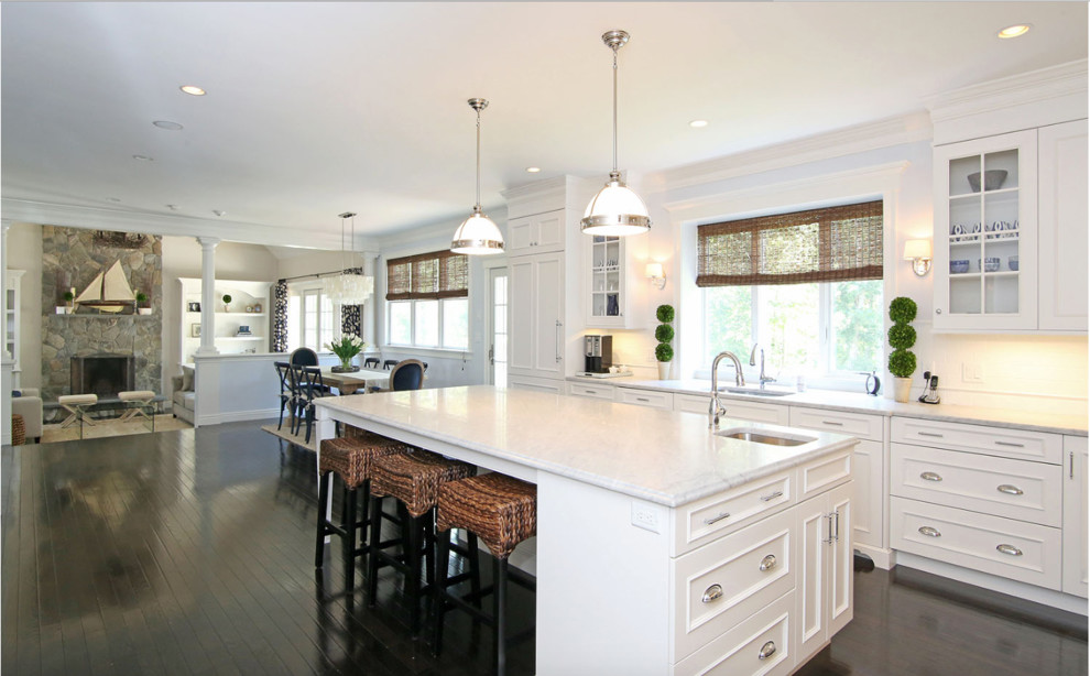 Large beach style l-shaped open plan kitchen in Boston with an undermount sink, flat-panel cabinets, white cabinets, marble benchtops, white splashback, subway tile splashback, panelled appliances, dark hardwood floors, with island and brown floor.
