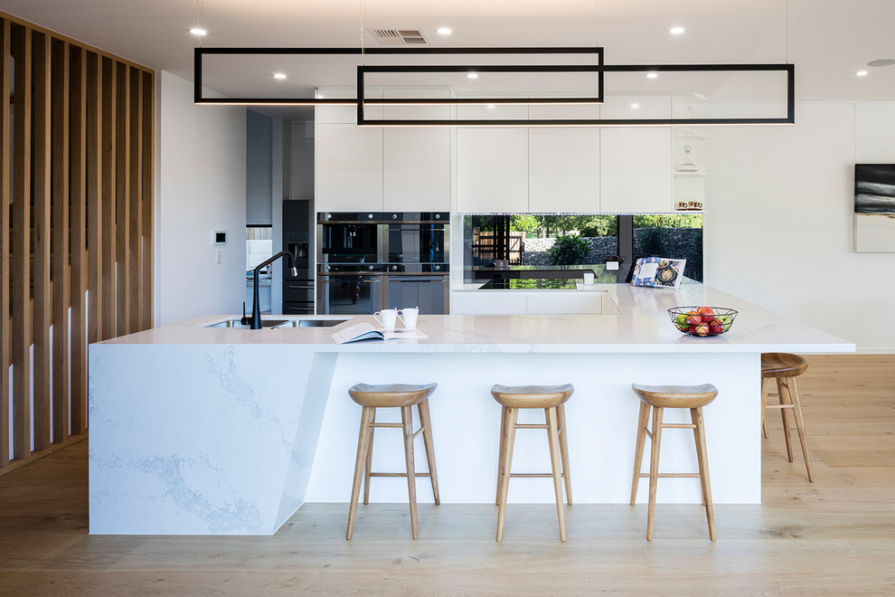 Photo of a mid-sized contemporary u-shaped open plan kitchen in Brisbane with a double-bowl sink, white cabinets, quartzite benchtops, metallic splashback, mirror splashback, black appliances, light hardwood floors, no island, beige floor, white benchtop and flat-panel cabinets.