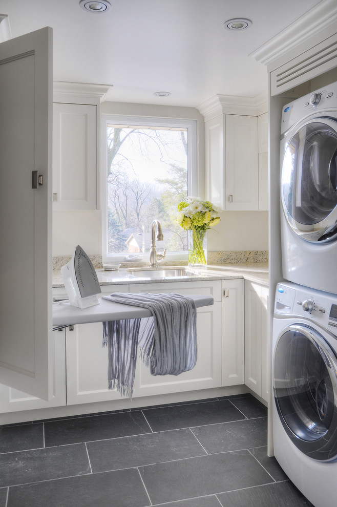 Photo of a mid-sized transitional u-shaped dedicated laundry room in Toronto with a stacked washer and dryer, white cabinets, an undermount sink, recessed-panel cabinets, granite benchtops, slate floors and beige benchtop.
